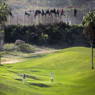 La photo de Jose Palazon montre deux joueurs de golf concentrés sur leur swing alors qu'une dizaine de migrants sont suspendus au sommet d'une grille à la frontière entre le Maroc et l'Espagne dans l'enclave de Melilla. [Jose Palazon]