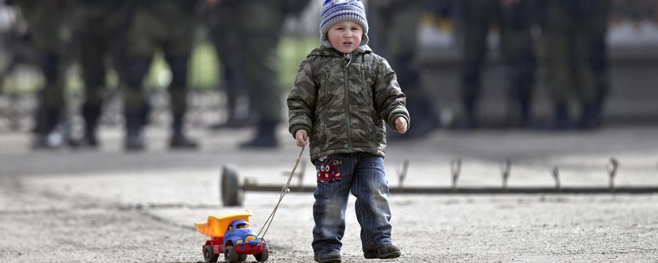 Enfant et son jouet aux abords d'une base militaire ukrainienne à Perevalne (Crimée), surveillée par des soldats pro-russes (arrière-plan). [Vadim Ghirda]