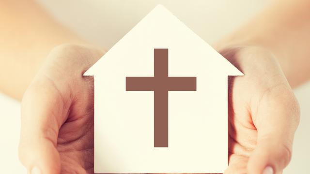 Des mains tenant une maison en papier avec une croix catholique. [Fotolia - Syda Productions]