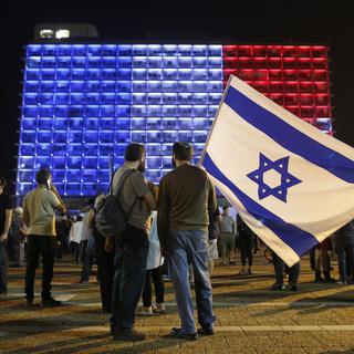 Un homme tient un drapeau d'Israël devant un bâtiment de Tel Aviv illuminé aux couleurs de la France. [EPA/Keystone - Abir Sultan]