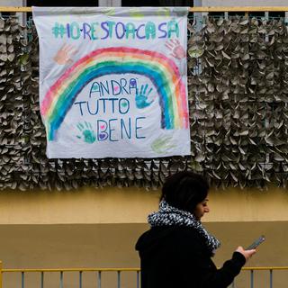 A woman passes by a sheet with the drawing of a rainbow and the inscriptions "Io resto a casa" (I stay at home) and "Andrà tutto bene" (Everything will be fine) made by a child displayed on a balcony in Pozzuoli, southern Italy, on March 13, 2020 as an initiative to reassure children and spread a message of trust and hope after the restrictions to combat the new Coronavirus, COVID-19. A government decree, after World Health Organisation (WHO) declared coronavirus a pandemic, ordered the closure of all commercial activities except for pharmacies and grocery stores and forces citizens to stay home. [CONTROLUCE /AFP - ELIANO IMPERATO]