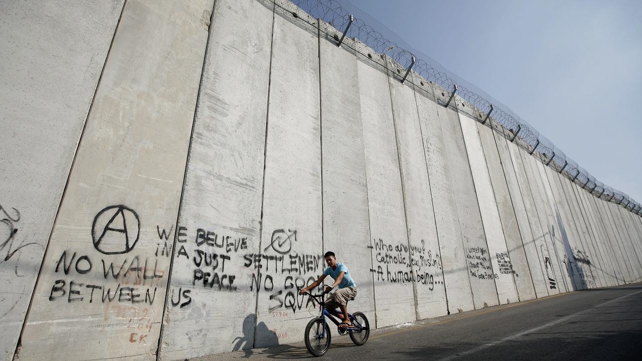 Un Palestinien sur son vélo à côté du mur séparant la Palestine d'Israël. [Keystone/EPA - Oliver Weiken]