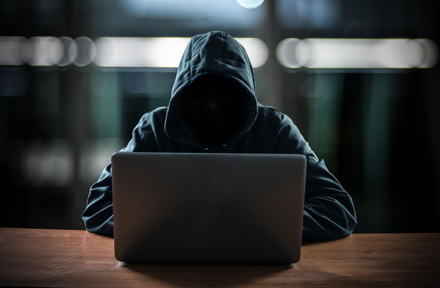 L'image cliché du hacker ou de la hackeuse: un personnage vêtu d'un pull à capuche, sans visage, penché sur un ordinateur dans l'obscurité. [Depositphotos - minervastock]