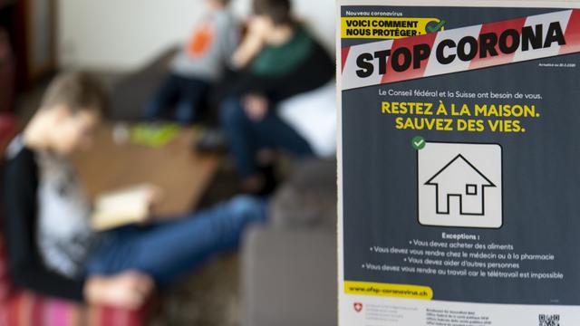 Une affichette de prévention concernant le nouveau Coronavirus (Covid-19) de l'office fédérale de la sante publique, OFSP, indiquant "Stop Corona, Restez a la maison. Sauvez des vies" photographiée dans le salon d'une maison ou trois enfants effectuent des activités lors de la pandémie du virus Coronavirus, (Covid-19) ce samedi 21 mars 2020 a Daillens. [KEYSTONE - Laurent Gillieron]