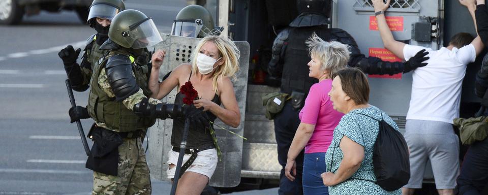 Une jeune femme armée d'un bouquet de fleurs aux prises avec un policier anti-émeute à Minsk, capitale de la Biélorussie, le 11 août 2020. Lorsque les Biélorusses ont protesté, le 9 août, contre ce qu'ils ont appelé une élection frauduleuse, la police a sévèrement réprimé les manifestations. Au pouvoir depuis 1994, le président Alexandre Loukachenko s'est efforcé de mettre un terme aux manifestations par de vagues promesses de réformes mêlées à des menaces, des convocations en justice et l'emprisonnement sélectif de militants de premier plan. [Keystone/AP]