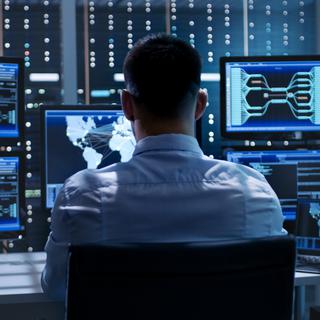 Homme de dos regardant des écrans d'ordinateur sur lesquels des données circulent. [Depositphotos - Gorodenkoff]