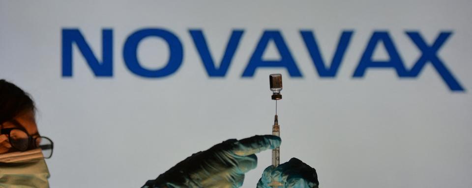 Une personne tient une seringue médicale et un flacon de vaccin devant le logo de Novavax. [AFP - Artur Widak / NurPhoto]