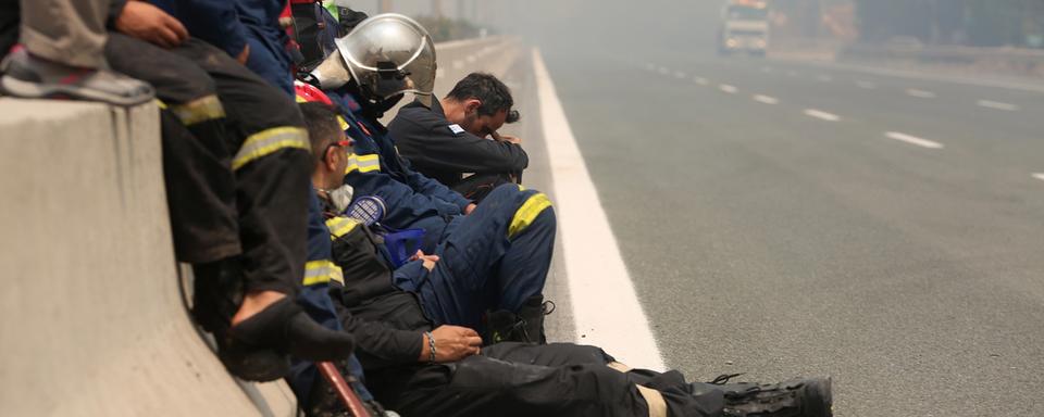 Vendredi 6 août: épuisés par une interminable lutte contre les incendies, des pompiers reprennent haleine sur le bas-côté de l'autoroute Athènes-Lamia. [Keystone/EPA - Alexander Beltes]