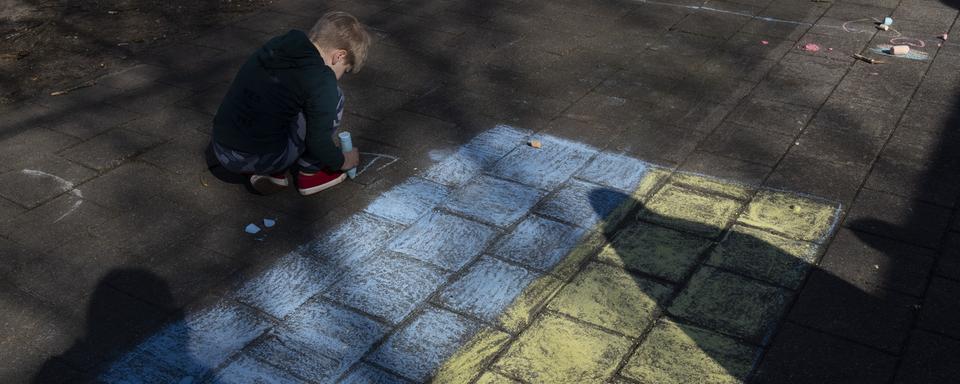 Un enfant dessine à la craie près d'un drapeau ukrainien lors de la visite du roi des Pays-Bas Willem-Alexander et de la reine Maxima dans un centre de réfugiés temporaire à Harskamp, aux Pays-Bas, jeudi 10 mars 2022. [AP Photo/KEYSTONE - Peter Dejong]