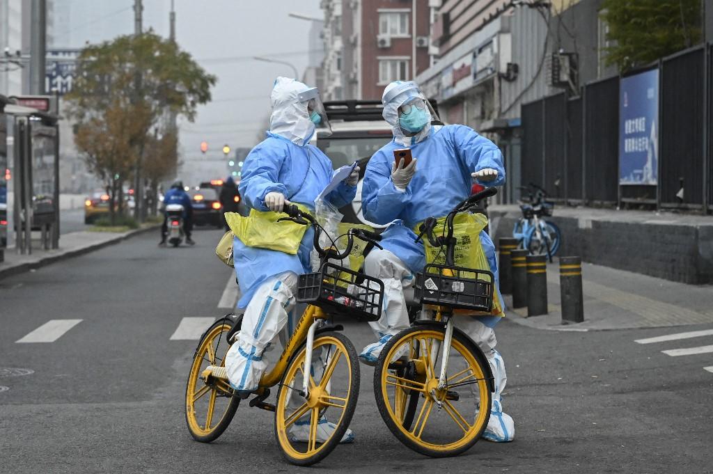 Des travailleurs sanitaire protégés des pieds à la tête se déplacent en vélo dans les rues de Pékin, où les cas de Covid ont atteint un record, le 24 novembre 2022. [AFP - Jade GAO]
