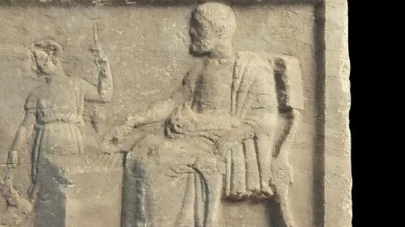 La scène représente un homme barbu et un garçon positionnés autour d’un ensemble de cinq lignes gravées sur un bloc. [Diachronic Museum of Larissa - Courtesy Larissa Museum]