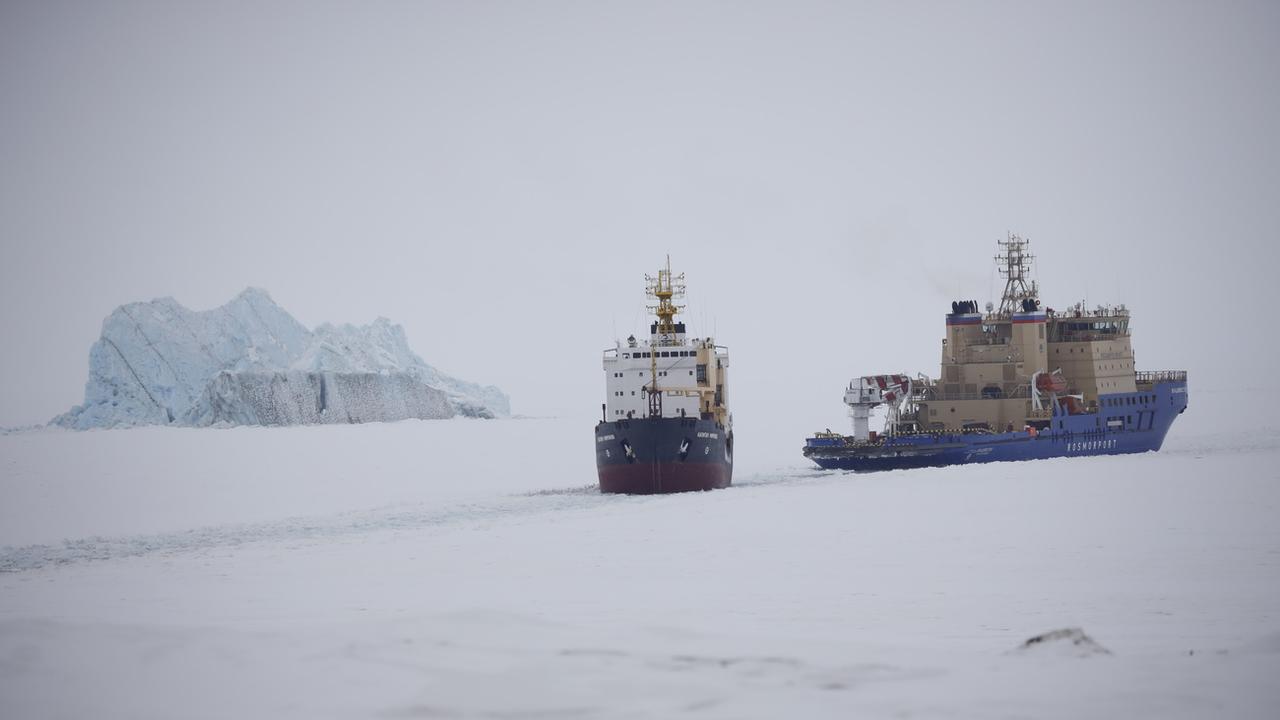 Un brise-glace trace la voie pour un cargo, avec un iceberg en arrière-plan, près d'un port sur l'île de la Terre d'Alexandra, près de Nagurskoye, en Russie. [KEYSTONE - Alexander Zemlianichenko / AP Photo]