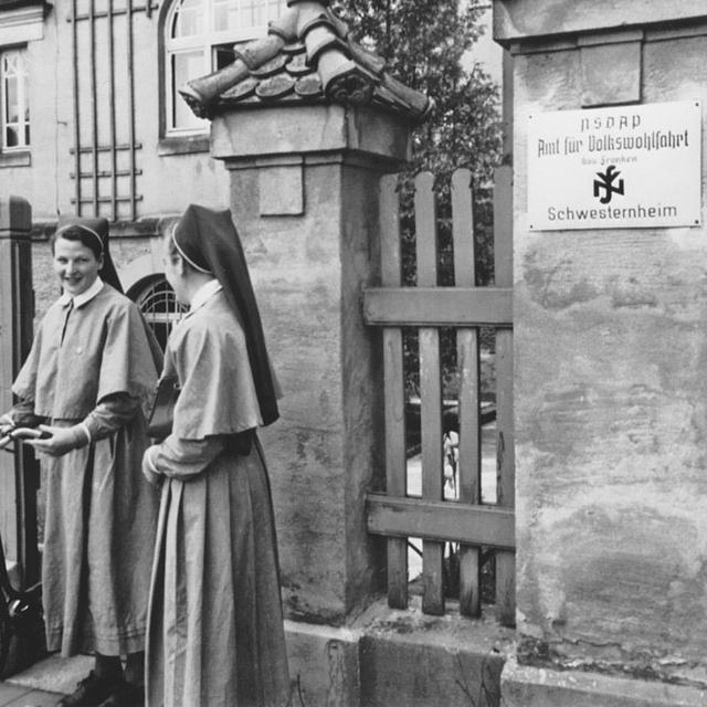 Deux soeurs infirmières à Erlangen (nord de la Bavière) en 1936. Sur la plaque à l'entrée de l'édifice on peut voir le sigle du NSDAP (Nationalsozialistische Deutsche Arbeiterpartei), l'acronyme allemand du parti national-socialiste, ou parti nazi, ainsi que la mention "Amt für Volkswohlfart", qui était le nom du secours populaire national-socialiste.