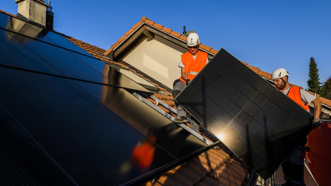 Deux installateurs implantent des panneaux solaires sur le toit d'une maison. [Keystone - Jean-Christophe Bott]