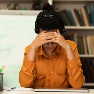 Une jeune femme assise à un bureau, portant un casque, se cachant le visage. [Depositphotos - Milkos]
