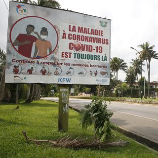Une femme passe devant un panneau affichant un message de sensibilisation au COVID-19 en français, indiquant "La maladie à coronavirus (COVID-19) est toujours là. Continuez à respecter les mesures de barrières", à Abidjan, Côte d'Ivoire, le 5 août 2021. [EPA/KEYSTONE - LEGNAN KOULAKOULA]