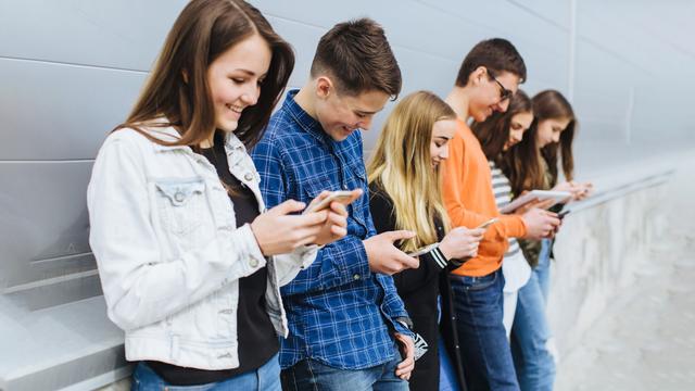 Six adolescents et adolescentes adossés à un mur consultent leur smartphone. [Depositphotos - anatoliycherkas]