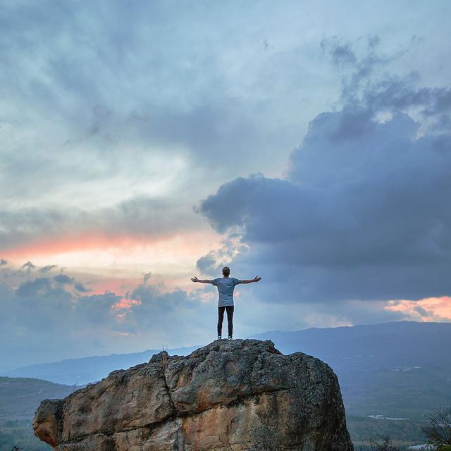 Un homme se tient debout, les bras ouverts, sur un rocher, face au paysage. [Unsplash - Joshua Earle]