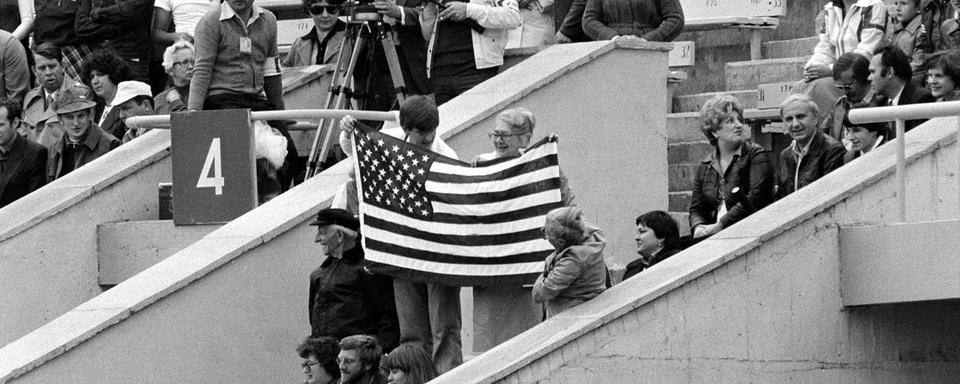 Trois spectateurs avec un drapeau américain dans les tribunes du stade Lénine de Moscou avant le début de la cérémonie d'ouverture des XXIIe Jeux olympiques d'été, le 19 juillet 1980. Cette année-là, soixante-deux nations décident d'un boycott des jeux initié par les États-Unis pour protester contre l'invasion soviétique de l'Afghanistan. En représailles, lors des JO de 1984 organisés à Los Angeles (USA), quatorze nations du bloc de l'Est ou sympathisantes décident de ne pas participer Jeux. [© KEYSTONE/AP Photo/Str]