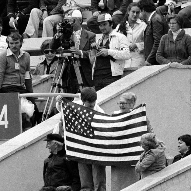 Trois spectateurs avec un drapeau américain dans les tribunes du stade Lénine de Moscou avant le début de la cérémonie d'ouverture des XXIIe Jeux olympiques d'été, le 19 juillet 1980. Cette année-là, soixante-deux nations décident d'un boycott des jeux initié par les États-Unis pour protester contre l'invasion soviétique de l'Afghanistan. En représailles, lors des JO de 1984 organisés à Los Angeles (USA), quatorze nations du bloc de l'Est ou sympathisantes décident de ne pas participer Jeux. [© KEYSTONE/AP Photo/Str]
