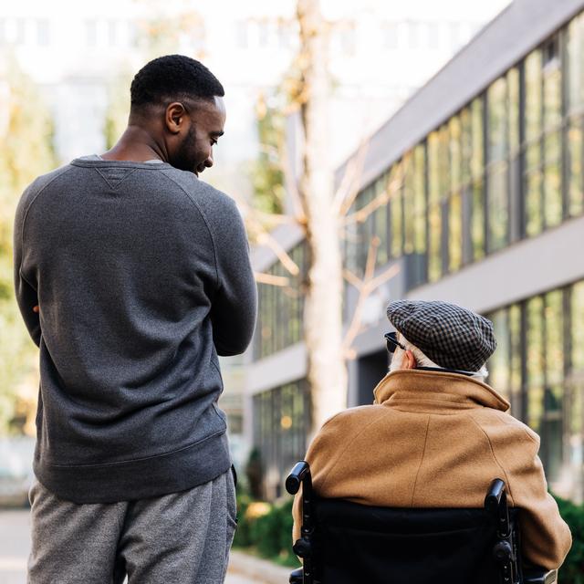 Un jeune homme et un sénior en chaise roulante se promènent dans la rue. [Depositphotos - Igor Vetushko]