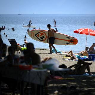 People cooling off in the shade of trees and a person carries his paddle board during warm weather on the shore of the Lake Geneva at the Vidy beach (plage de Vidy) in Lausanne, Switzerland, Sunday, June 19, 2022. People flocked to parks and pools across Western Europe on Sunday for a bit of respite from an early heat wave that saw the mercury rise above 40 Cs (104 F) in France and Spain, and highs of 38 C (100.4 F) in Germany and 36 C in Switzerland. (KEYSTONE/Laurent Gillieron) [Keystone - Laurent Gillieron]