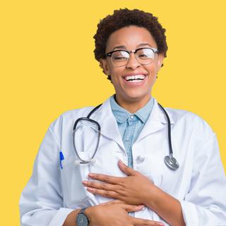 Jeune femme médecin afro-américaine portant un manteau médical sur fond isolé Sourire et rire fort à haute voix parce que drôle blague folle. Bonne expression. [Depositphotos - ©Krakenimages.com]