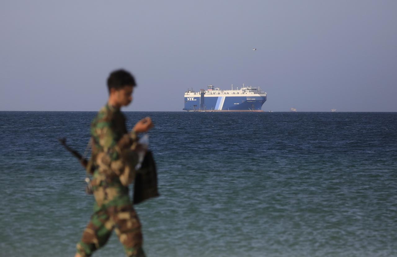 Un combattant du groupe rebelle marche sur la plage, avec en arrière fond le Galaxy Leader. Les Houthis se sont emparé du cargo le 19 novembre 2023. [Keystone - Yahya Arhab/EPA]