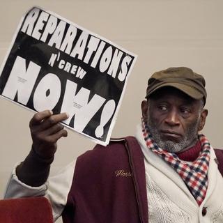 Un homme brandit une pancarte lors d'une réunion du groupe de travail chargé d'étudier et d'élaborer des propositions de réparation pour les Afro-Américains à Oakland, en Californie, le mercredi 14 décembre 2022. [Keystone - AP Photo/Jeff Chiu]