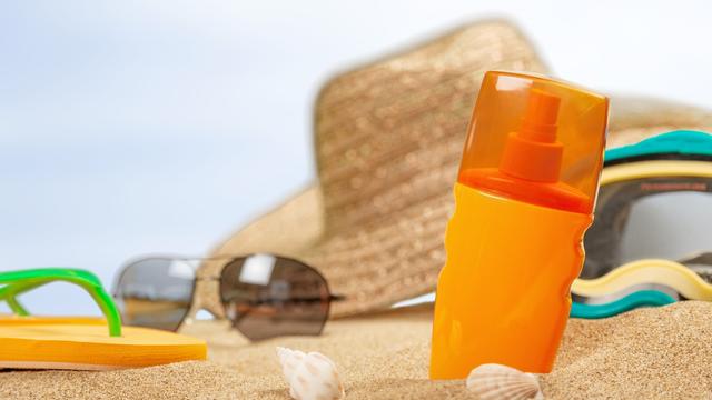 Sur une plage, un chapeau, des lunettes, des tongs et un flacon de crème solaire sont posés sur le sable. [Depositphotos - Billiondigital]