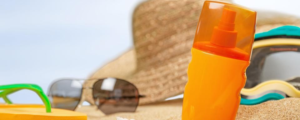 Sur une plage, un chapeau, des lunettes, des tongs et un flacon de crème solaire sont posés sur le sable. [Depositphotos - Billiondigital]