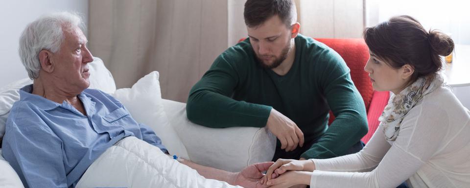 Un homme dans un lit d'hôpital accompagné de deux personnes plus jeunes. [Depositphotos - photographee.eu]