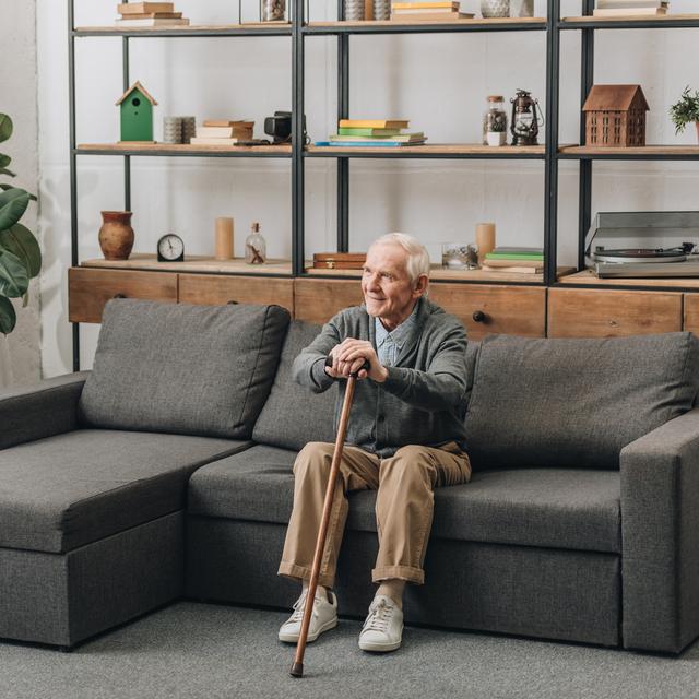 Heureux homme âgé souriant et tenant canne à pied tout en étant assis sur le canapé. [Depositphotos - ©AndrewLozovyi]
