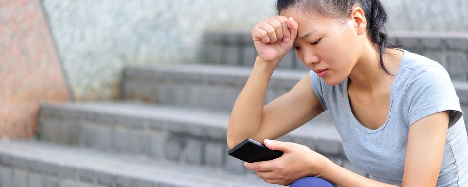 Une femme assise sur des escaliers dans la rue regarde son téléphone d'un air anxieux. [Depositphotos - Izf]