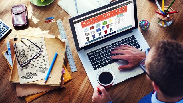 Un homme assis à son bureau consulte un site de vente de vêtements sur son ordinateur, café à la main. [Depositphotos - Rawpixel]