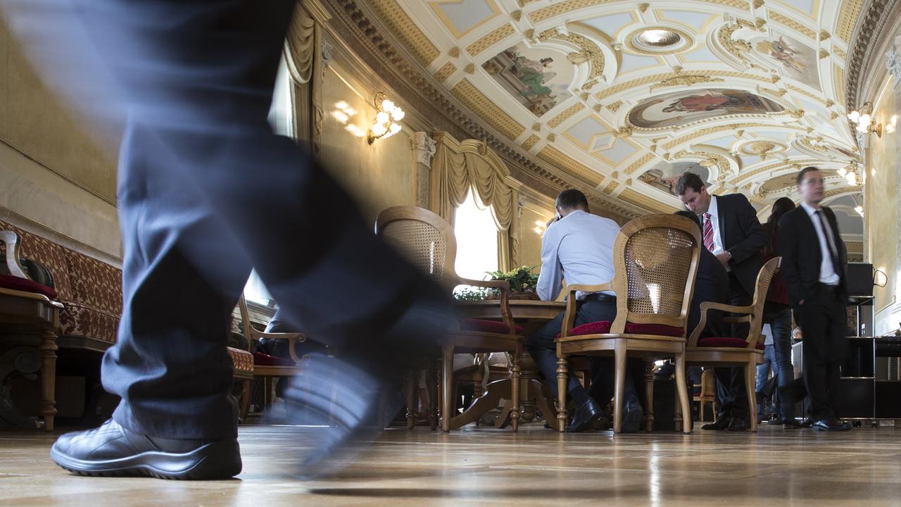 Vue de la salle des pas perdus du Parlement fédéral à Berne. Au premier plan, les pieds d'un homme qui marche. Au second plan, plusieurs personnes en costume sont en discussion. [Keystone - Peter Klaunzer]