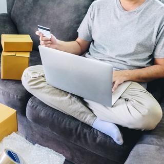 Un homme est assis sur un canapé avec un ordinateur sur les genoux. Il est entouré de cartons et tient une carte de crédit à la main. [Depositphotos - Freedomtumz]