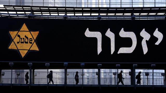 Des personnes marchent le long d'un pont éclairé par un panneau d'affichage montrant une étoile de David jaune qui porte l'inscription "Jude" (juif en allemand) et le mot "Remember" (se souvenir) en hébreu, lors de la journée annuelle de commémoration de la Shoah à Ramat Gan, en Israël, mardi 18 avril 2023. [Keystone - Oded Balilty / AP Photo]