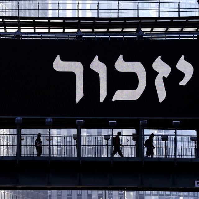 Des personnes marchent le long d'un pont éclairé par un panneau d'affichage montrant une étoile de David jaune qui porte l'inscription "Jude" (juif en allemand) et le mot "Remember" (se souvenir) en hébreu, lors de la journée annuelle de commémoration de la Shoah à Ramat Gan, en Israël, mardi 18 avril 2023. [Keystone - Oded Balilty / AP Photo]
