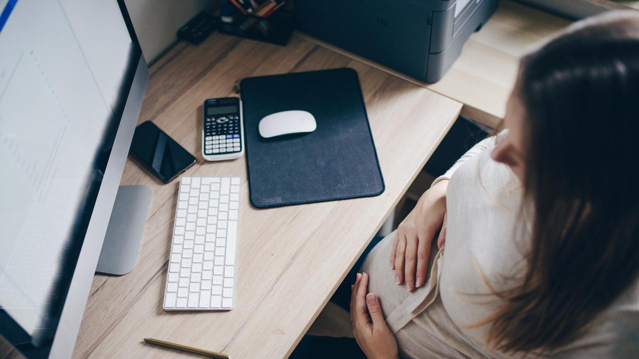 Une femme enceinte est assise à un bureau. Elle regarde l'écran de son ordinateur tout en se tenant le ventre avec ses mains. [Depositphotos - Samanyuk]
