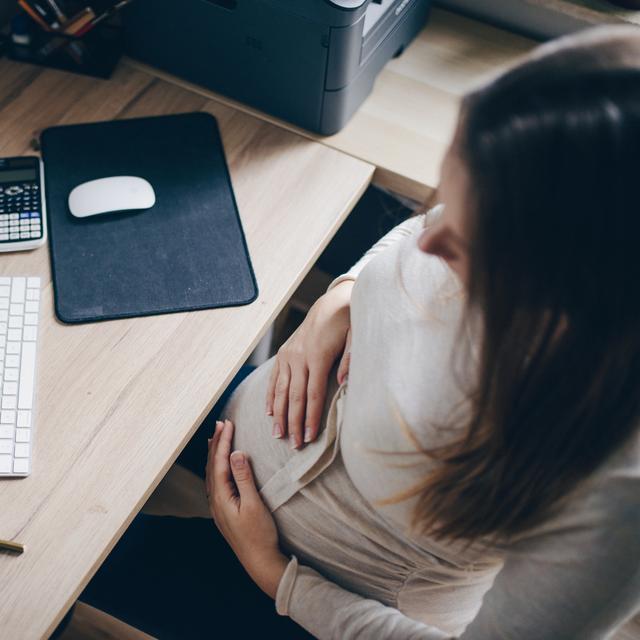 Une femme enceinte est assise à un bureau. Elle regarde l'écran de son ordinateur tout en se tenant le ventre avec ses mains. [Depositphotos - Samanyuk]