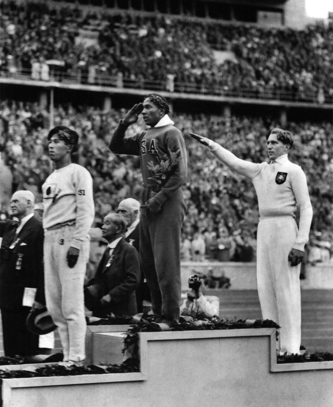 L'Américain Jesse Owens (au centre), salue lors de la présentation de sa médaille d'or pour le saut en longueur, le 11 août 1936, après avoir battu l'Allemand Lutz Long (à droite), lors des Jeux olympiques d'été de 1936 à Berlin. Le Japonais Naoto Tajima (à gauche), s'est classé troisième. Jesse Owens a triomphé dans les compétitions d'athlétisme, remportant quatre médailles d'or dans les courses de 100 et 200 mètres, le saut en longueur et le relais 400 mètres. Il est le premier athlète à remporter quatre médailles d'or aux Jeux Olympiques. [KEYSTONE - STR]