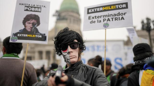 A masked anti-government protester aims a toy gun during a demonstration outside Congress where lawmakers debate a reform bill promoted by President Javier Milei in Buenos Aires, Argentina, Wednesday, June 12, 2024. The sign at right reads in Spanish "Exterminator of the homeland." [AP Photo/Keystone - Natacha Pisarenko]