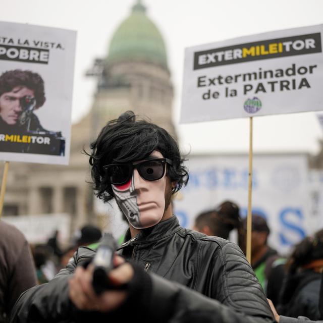 A masked anti-government protester aims a toy gun during a demonstration outside Congress where lawmakers debate a reform bill promoted by President Javier Milei in Buenos Aires, Argentina, Wednesday, June 12, 2024. The sign at right reads in Spanish "Exterminator of the homeland." [AP Photo/Keystone - Natacha Pisarenko]