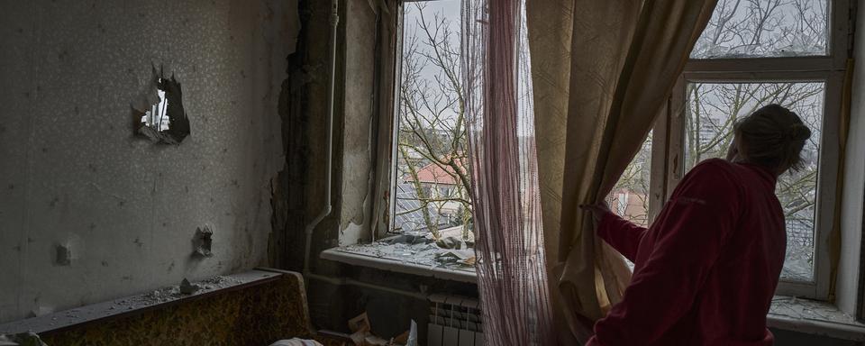 Une femme ukrainienne regarde à travers la fenêtre de son appartement bombardé. [Keystone/AP Photo - LIBKOS]