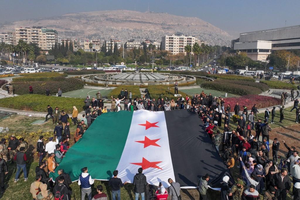 Des personnes tiennent un grand drapeau de l'opposition syrienne sur la place des Omeyyades, à Damas, le 9 décembre 2024. [AFP - OMAR HAJ KADOUR]