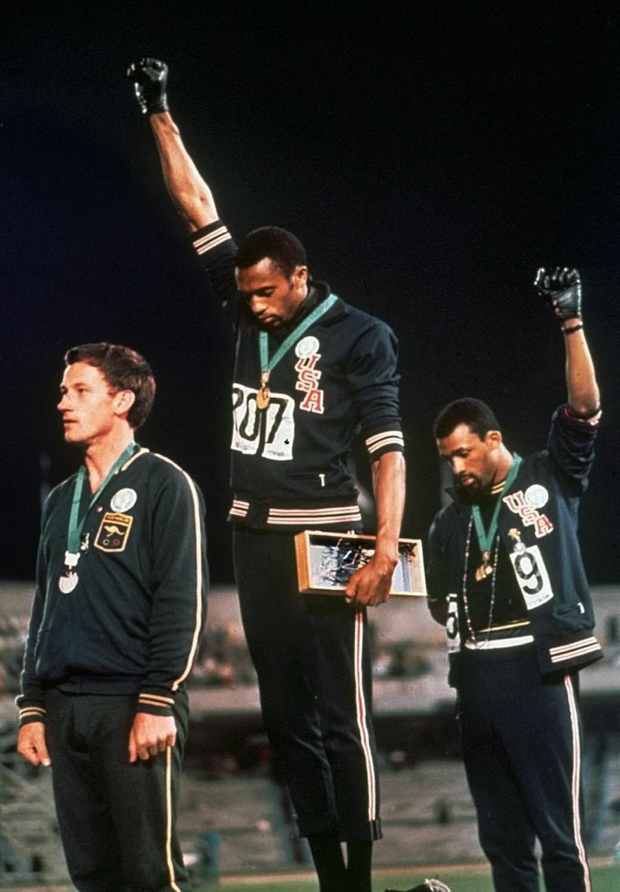 Tommie Smith, au centre, et John Carlos, deux athlètes américains, tendent leurs mains gantées vers le ciel en signe de protestation contre la ségrégation raciale aux USA et regardent vers le bas pendant l'interprétation de l'hymne national américain, après que Tommie Smith ait reçu l'or et John Carlos le bronze pour la course de 200 mètres aux Jeux olympiques de Mexico, le 16 octobre 1968. [KEYSTONE - ANONYMOUS]