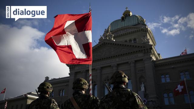 Trois soldats de l'armée suisse en rang devant le Palais fédéral à Berne. L'un d'entre eux tient un drapeau à croix blanche. [KEYSTONE - ANTHONY ANEX]