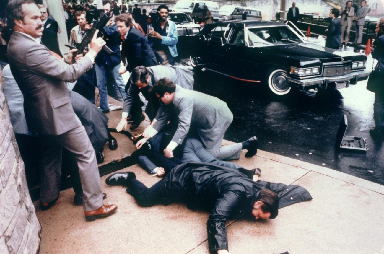 Photo des tumultes quelques secondes après l'attentat contre Ronald Reagan. Des membres du personnel entourant le président, aussi touchés par des tirs, sont au sol. [Keystone - EPA, WHITE HOUSE]