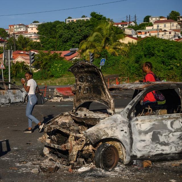 Des piétons passent derrière une voiture brûlée suite aux récentes manifestations sur le coût de la vie, à Fort-de-France, Martinique, le 23 septembre 2024. [AFP - ED JONES]